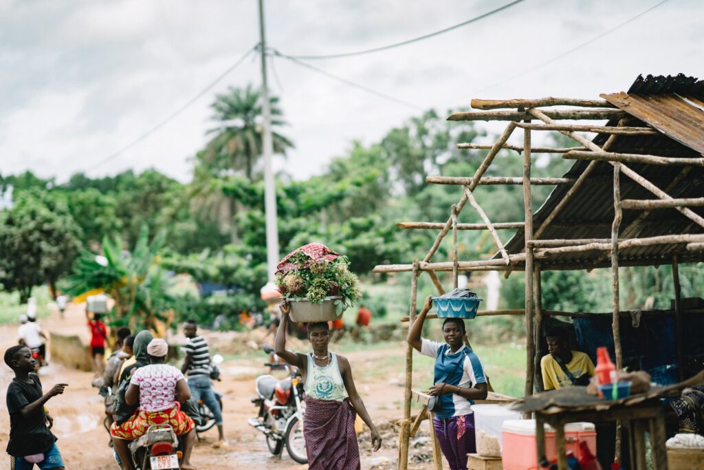 People on a dirt road in a rural community, carrying baskets of fruit or riding motorcycles.
